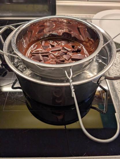 Ein Topf mit kochendem Wasser, darin eine Edelstahlschüssel mit schmelzender Schokolade, die durch einen einfachen Draht-Kleiderbügel über Wasser gehalten wird.