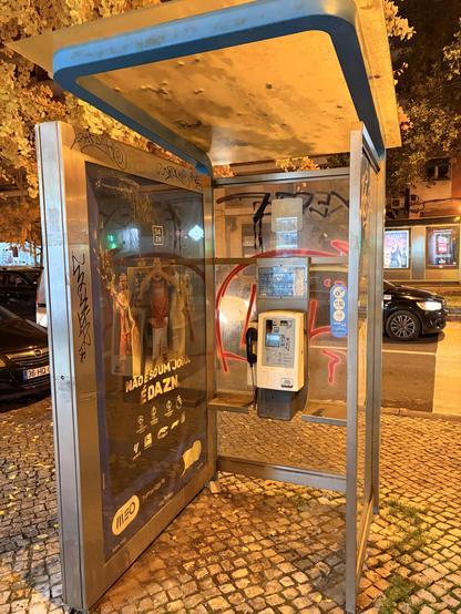 A Portuguese pay phone with glass walls. The booth is open on one side, and graffiti has been unceremoniously sprayed on the outer walls.
