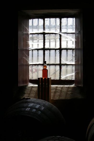 Das Bild zeigt ein Innenarrangement vor einem Fenster. Das Fenster besteht aus mehreren quadratischen Fenstern, die durch dunkle Balken geteilt sind und im unteren Bereich durch ein feines Spinnennetz überzogen sind. Im Fenster befindet sich eine orangefarbene Flasche auf einem schmalen, dunklen Sockel. Die Flasche befindet sich im Zentrum des Bildes. Im Vordergrund befindet sich ein großer, runder, dunkler Gegenstand, der nur teilweise im Bild zu sehen ist. Das Arrangement ist stark von Schatten umgeben und wirkt stimmungsvoll.