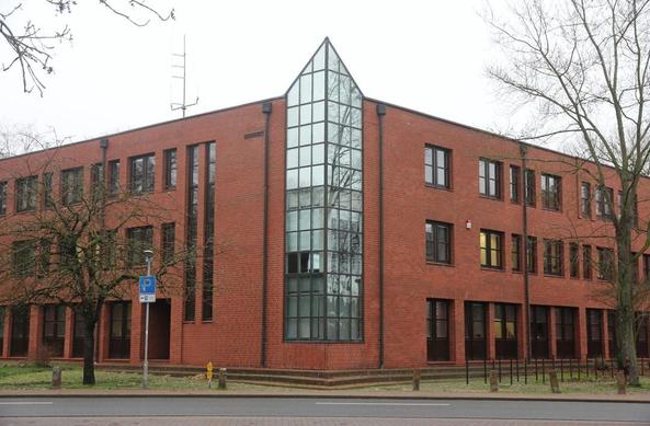 Dreigeschossiges Bürogebäude mit roter Fassade und großen Glasfenstern, davor eine Straße und Bäume.
