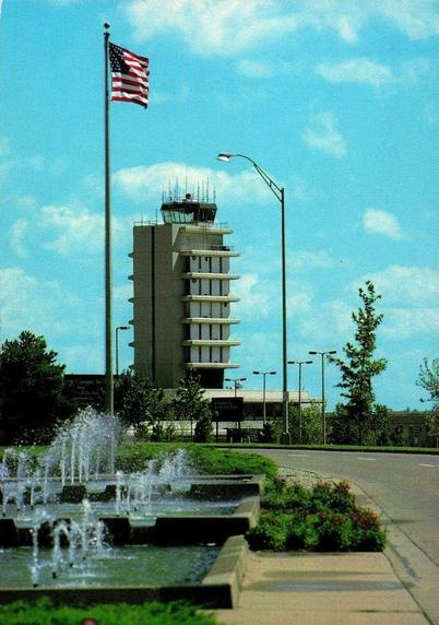 This postcard captures the dramatic approach to Kent County International Airport, where visitors were greeted by a broad reflecting fountain, manicured landscaping, and the airport’s striking mid-century control tower rising in the distance. The tower, with its stacked concrete balconies and projecting cab, served as both an operational hub and an architectural landmark for the region, symbolizing Grand Rapids’ aspirations for modern air travel. The scene reflects a period when airports emphasized civic presence and ceremonial entryways, using fountains, open plazas, and flagpoles to convey a sense of welcome and national pride.
