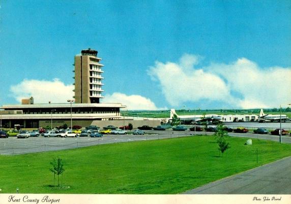 This image shows the airport shortly after its major 1960s expansion, featuring the newly built terminal with its broad horizontal lines, deep overhangs, and the modernist control tower rising above it. In the background, propliner-era aircraft are parked on the apron, illustrating the transitional moment when turboprops and piston aircraft still dominated the field just before the full arrival of commercial jets. The foreground parking lot—organized in neat rows—reflects the era’s growing reliance on the automobile and the airport’s shift toward serving a much broader traveling public. Set against open fields and wide skies, the scene captures Kent County Airport at a moment when air travel was becoming central to regional economic identity.
