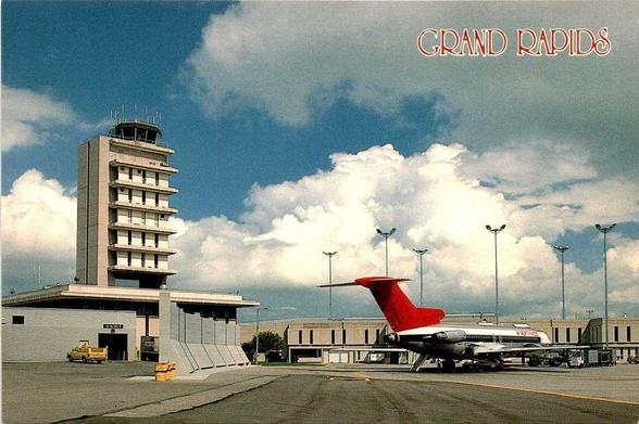 This view highlights the busy flightline at Kent County International Airport during the peak of the jet age, anchored by the tall control tower and the terminal complex spreading low and wide beneath it. A Northwest Airlines Boeing 727—once the workhorse of domestic trunk routes—sits on the ramp undergoing servicing. Light towers, baggage carts, and ground-handling equipment convey the airport’s role as a regional gateway connecting West Michigan to major national hubs. The tower’s distinctive vertical massing contrasts with the low-slung terminal buildings, a design typical of mid-century airport expansions that prioritized visibility for controllers and efficient movement for passengers.