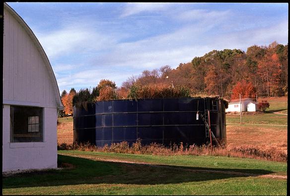 A black short cylinder filled with grass sits in a field next to a barn, which is partly visible at left.