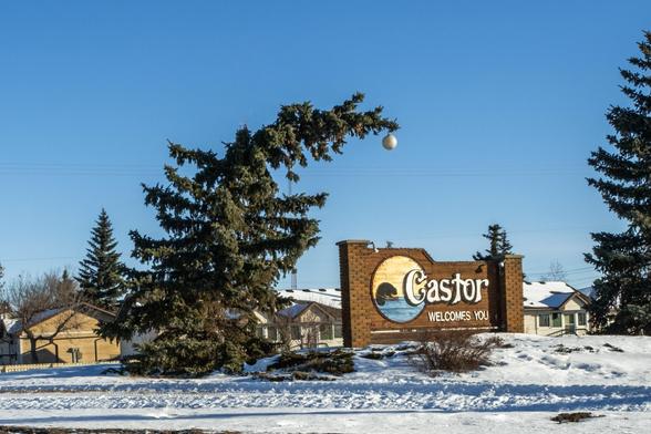 A photo of the "Castor Welcomes You" sign outside of Castor, Alberta, with a spruce tree leaning over to make it look like a Charlie Brown Christmas tree, adorned with the classic big ball on the end.