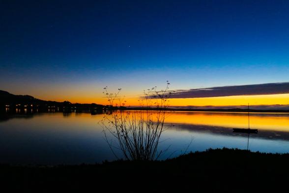A wide shot depicts a calm body of water, likely a lake or bay, at dusk or dawn. The sky transitions from a deep blue at the top to a vibrant orange and yellow band near the horizon. Silhouetted against this colorful backdrop are landmasses, possibly hills or a distant shoreline. In the foreground, a patch of tall, dry grasses extends from the lower edge of the frame towards the water, creating a dark, plant-like outline. A small boat is visible as a dark shape on the water's surface, reflecting the sky's colors.