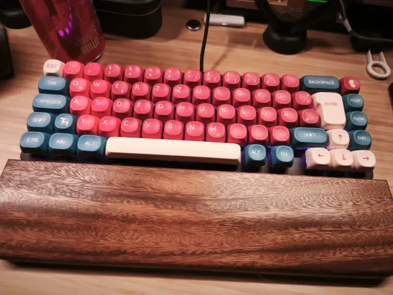 eine kleine 65% Tastatur liegt auf einem Holztisch. Davor eine Handballenauflage aus dunklerem Holz.

Die Tasten sind leicht abgerundet und mittig in rot-tönen und drum herum mit blauen Akzenten.