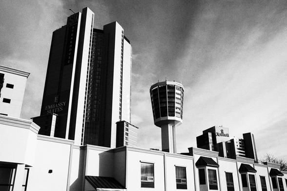 A high-contrast black-and-white photograph showing tall hotel towers rising above lower foreground buildings. The Embassy Suites tower dominates the left side, while the Skylon-style observation tower stands prominently to the right. Dramatic cloud patterns sweep across the sky, adding texture above the stark architectural forms.