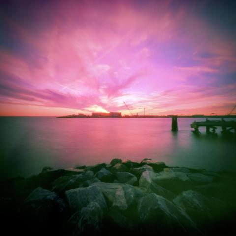 Sunset over water with an island in the background, rocks in the foreground, and a decaying pier off to one side.