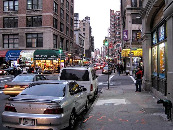 Katunäkymä New Yorkin Chinatownissa. Kadun varressa on pysäköityjä autoja ja jalkakäytävällä kulkee joitain jalankulkijoita.