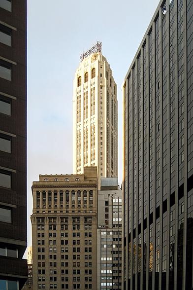 Korkeiden talojen takana nousee yksinäinen pilvenpiirtäjä taivasta kohti. Etualalla olevat talot ovat varjossa ja takana korkeuksiin nouseva pilvenpiirtäjä on auringon kirkkaasti valaisema.