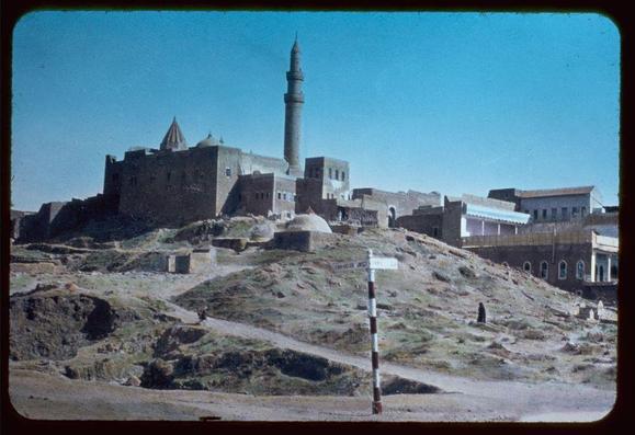 This image depicts a historical site, likely of religious significance, set atop an elevated terrain. A prominent feature is the mosque with its towering minaret and domes, which suggests Islamic architecture. The complex appears to be surrounded by fortified walls, indicative of either defensive purposes or status symbolism during its time.

The surrounding landscape shows arid conditions, characterized by sparse vegetation and rocky ground. There are several people visible in the scene: one person is walking along a dirt path leading towards the mosque, while others appear further down the hillside, which has a rough terrain with uneven paths cutting through it.

A signpost stands at the forefront of the photo on what looks like a roadside or pathway that leads to the site. The overall atmosphere evokes a sense of historical significance and cultural heritage amidst an arid landscape, potentially indicative of locations in regions such as Mesopotamia during its ancient history.