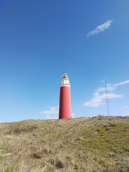 A red lighthouse stands on a grassy slope under a clear blue sky with a single wispy cloud. The lighthouse, painted with white accents around the windows, sports a classic conical structure topped with a white lantern room and gallery. To its right, a thin metallic tower appears in the distance.
