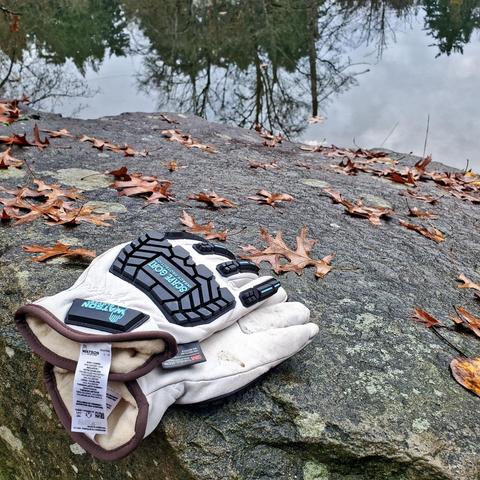 A pair of white and black work gloves, branded "WATSON GLOVES", rest on a textured, wet rock surface strewn with fallen brown leaves. A calm, water body with reflected trees and overcast sky is in the blurred background.