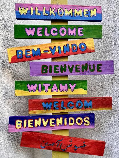 Vertical arrangement of multicolored wooden planks against a light gray wall, each painted in vibrant colors with the word "welcome" in different languages. The languages include German, English, Portuguese, French, Polish, and Spanish, displayed in a variety of bright color backgrounds. The text "Khosh Amadid" in Persian is also included at the bottom plank in a cursive blue script.