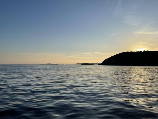 Sunset over a calm sea with gentle ripples, featuring a silhouette of a forested coastline on the right and distant islands on the horizon under a clear sky. The sun, partially visible, casts a warm glow near the top right corner.