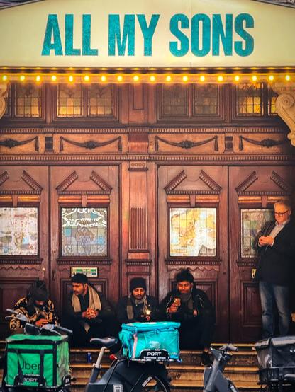 On the steps outside a theatre, four guys in their late twenties to early thirties are sitting waiting to do their next food bike delivery. Their bikes can be seen in front of them, at the bottom of the picture. They are dark-skinned with black hair, and are perhaps Asian. To their right, standing at the top of the steps, is a fair-haired much older white guy, looking at his phone.