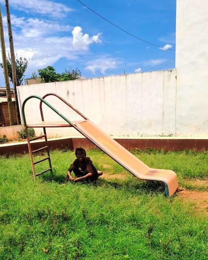 Two children playing under a rusty metal slide in a grassy area. The slide, with its green metal handles and ladder, stands against a backdrop of a white wall and a clear blue sky. The child in front is smiling and crouching, while the second child is mostly hidden behind them.