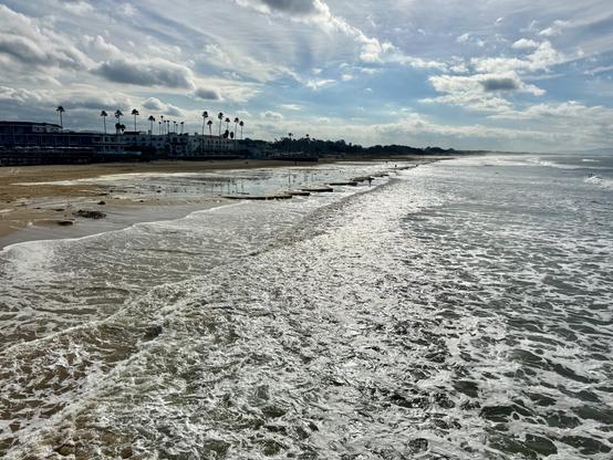 A shot of the waves rolling into the coastline of Pismo Beach, California. The shot is looking into the sun, so the water looks silver due to the reflection. Along the coast are tall palm trees that rise above everything else. The sky is fairly cloudy, but the sun was shining on the waves for this shot.