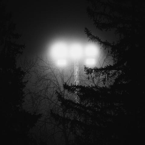 Light after dark

A greyscale monochrome square photograph of a large array of flood lights, on top of a slender tower.

the tower is partly obscure and the lights framed by a large spruce tree filling the right edge of the photo, silhouetted by the bright lights.

Another spruce silhouette fills the left edge.

Just behind and to the left of the light tower stands a deciduous tree, barren of leaves, standing out in the foggy atmosphere which is partly lit by the lights.

The uppermost part of the photo reveals the dark sky.