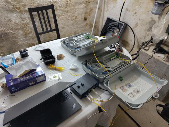 Ein Tisch im Keller mit einem Laptop, einem Banana Pi R4, zwei offenen Outdoor-Spleißboxen, sowie diverses Glasfaserinstallationsequipment. Alles fertig verkabelt zur Messung.