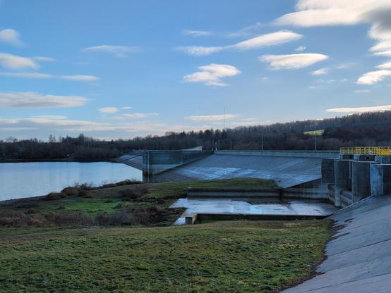 A view from the arc of a dam, towards its further part. To the left, some meadows and then the blue water of the lake. In a short distance there's the first floodgate, located over the current water level, with concrete bottom shining from the moisture and a bunch of round concrete elevations extending from the dam, with bright yellow railings on top. Further away the dam starts reaching the water level, and another floodgate appears, entirely concrete this time. A forest in the distance, to the right it gets hilly. The sky is blue with a few white clouds.