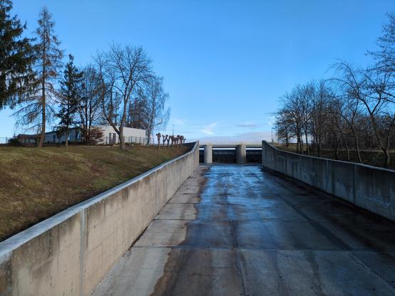 A view from a bridge in the middle of the exterior side of the floodgate. In the foreground, a wide and deep concrete canal for the water, empty right now. Except for a strip to the left, the bottom is brownish from past water, still shining with moisture. Further away, the floodgate itself on two rounded concrete columns. The walkway on top is surrounded by yellow railings. Behind it, steel barriers for the water. To the right side, trees are growing over the canal. To the left side, they've been cut to stumps.