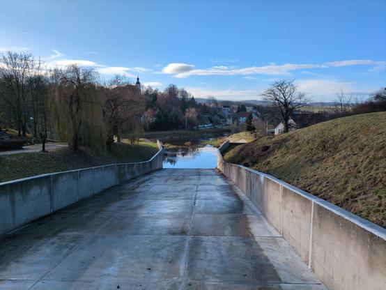 A view to the other side. The floodgate canal enters a river going left to right. There are some clumps of plants standing in the water near the outset. There's a town around the river, with hilly terrain. There are some houses and trees. To the left, a church tower emerges above, on a hill.