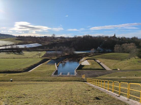 A view over the other floodgate, from up on the dam. This time the canal emerges much lower, and is filled with water. It joins the dam underground, it's covered with earth on top, with short grass on top. To the right, there are stairs going down (with yellow railing). Above the canal there's a concrete walkway going right, with dark metal railings (not yellow). To the sides, the river connects to additional concrete canals, that turn towards the dam in straight edges, then turn back parallel to it. The river turns right. To the right of it, there are green meadows. To the left, additionally plowed fields, some of them still covered by snow. Further away, some brownish trees in shadows. The church tower is also visible here in the distance.