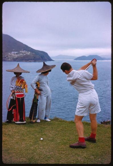 A man swinging a golf club, aiming towards the water with two people wearing traditional hats looking on and holding onto their golf bags. The background shows blue waters and islands in distance under cloudy skies. A white ball is visible near him as he plays his shot from grassy ground beside an ocean or sea.