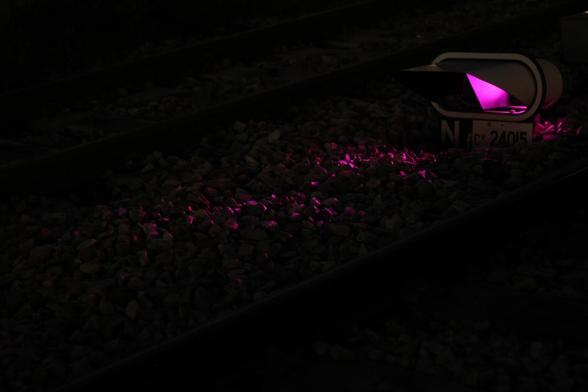 Photo, la nuit, d'une lumière de signalisation ferroviaire violette située à ras de ballast entre deux voies, et dont la lumière se reflète légèrement sur le ballast.