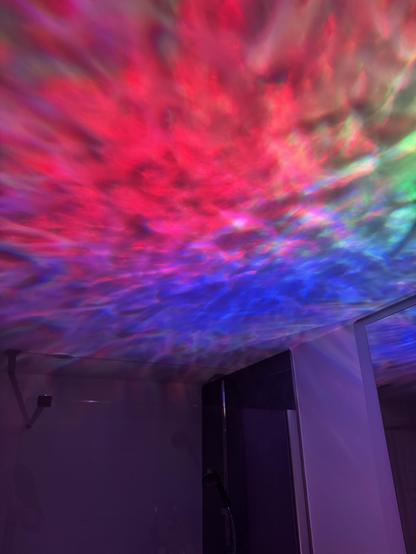 A small room with a shower and a glass wall with colourful lights projected onto the ceiling