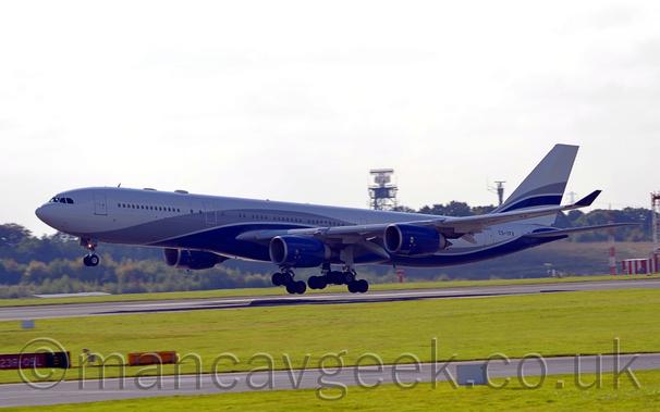 Side view of a 4 engined jet airliner flying from right to left at a very low altitude, just a couple of metres above a black runway, with flaps extended from the rear of the wing, undercarriage lowered, and the nose raised slightly, suggesting it is about to land.
The plane is mostly white, with thick grey and dark blue stripes coming from below the forward fuselage, sweeping up over the wings, and back down under the rear fuselage, the grey stripe getting narrower as it moves rearward.
A similar stripe runs across the tail.
The engine pods under the wings are dark blue, as are the up-turned wing tips.
The black registration "CS-TFX" is on the upper rear fuselage, under the tail.
Green grass fills the foreground, with a grey taxiway cutting through it from one side of the frame to the other.
Trees line the side of a low hill in the background, with a small radar tower at the top visible over the top of the planes body, under bright but hazy sky.