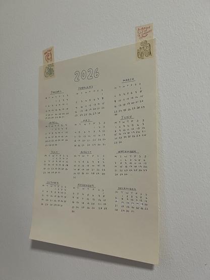 piece of paper with handwritten months and days. at the top it says 2026. it is held on the wall by two stripes of washi tape with cute animal illustrations.