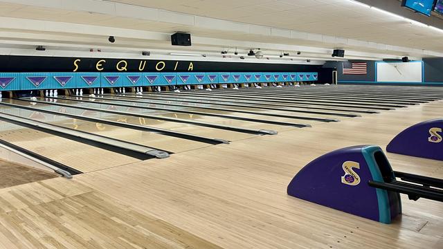 Bowling lanes stretch across a spacious alley. "SEQUOIA" letters are visible above the lanes. Bowling pins are set at the end of each lane, with American flag on the wall.