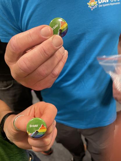 Zwei Buttons werden von zwei verschiedenen Menschenhänden in die Kamera gehalten. Man erkennt noch einen Teil des Aufdrucks auf dem türkis-blauen T-Shirt des einen: „DAV GOC“, „Der queere Alpenverein“, und das DAV Logo „Edelweiß“ in der Regenbogen Variante.
Auf dem Button erkennt man ein Alpenpanorama in den Farben der progressiv Pride Flagge auf grünem Hintergrund und die Buchstaben DAV in weiß.