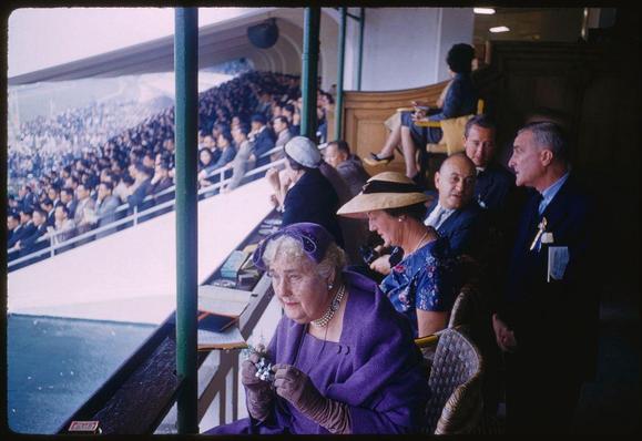 This image is a color transparency photograph taken in 1959 by Toni Frissell, known for her work as an American fashion and sports photographer. The scene captures the essence of Hong Kong during that time period, showcasing its blend of British colonial influences.
The main subject appears to be seated at an event or venue with spectators around them. They are dressed in elegant attire, including a woman wearing a purple coat adorned with jewelry, pearls, and gloves, which suggests formality and sophistication often associated with high-profile social gatherings during the mid-20th century. The surroundings hint at a crowded stadium setting where people gather to watch an event or performance.
This photograph is part of Toni Frissell's "Choice + Selects" collection, featuring various black-and-white and color images captured in different locations, including Hong Kong (marked as x5826).