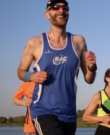 A lanky guy with a big goofy grin wearing sunglasses and a Rushcliffe Athletic Club vest. He’s bathed in a lovely warm evening sunshine glow and is running around Holmepierrepont Lake as part of a summer league club running event.