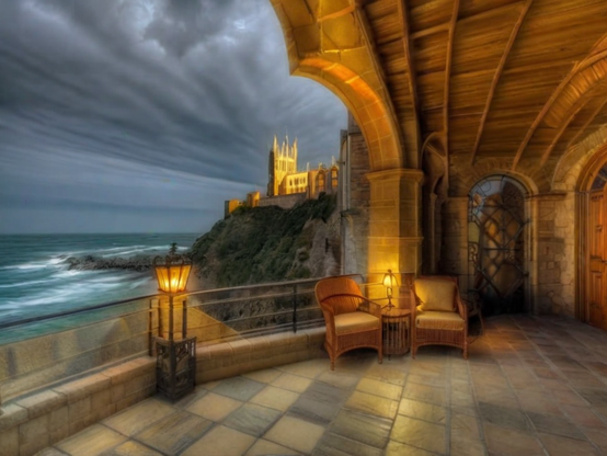 A warmly lit stone terrace with wicker chairs, overlooking stormy seas, opens to a very long castle running along coastal cliffs to a tower with spires glowing against the dramatic dusk sky.