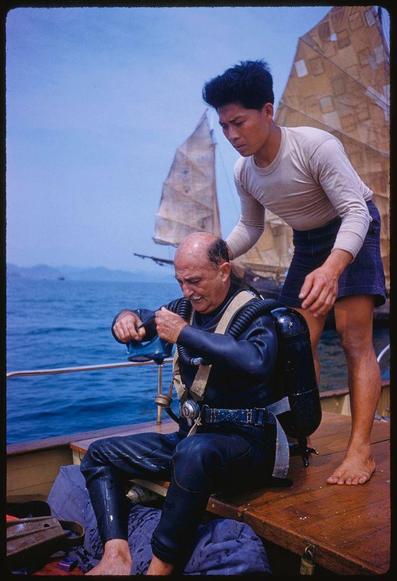 two men wearing black wetsuits and gear on a boat with other boats in the background near buildings. one is seated, while another stands over him helping him put something into his mouth that has tubing attached to it; he's adjusting or preparing for some kind of apparatus connected by tubes to an air tank strapped across chest
