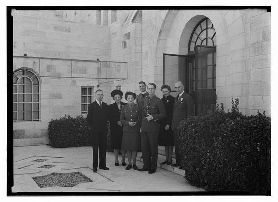 A black and white photograph showing a group of eight people, presumably from an earlier era given the fashion styles. They are standing on steps in front of what appears to be a significant building with large stone walls and arched windows. The architecture suggests it could be a governmental or institutional setting. Among them is someone wearing military attire, suggesting they may have served during World War II, which coincides with their age range from the early 1940s as per the date provided in the alt text information. They are standing on concrete steps leading up to an open door of this building, and there's a bush visible beside them. Their attire is formal for men wearing suits or uniforms, while women wear dresses and some have accessories such as hats or fur coats. The atmosphere seems solemn yet respectful given their posture and expressions.