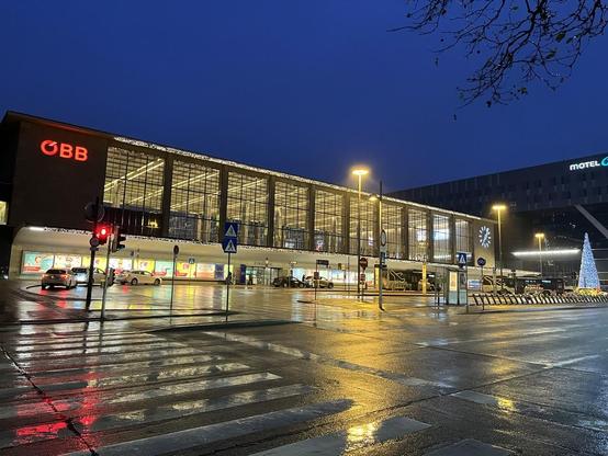 Der Westbahnhof, ein kastiger Nachkriegsbau mit einer hohen Fensterfront über dem Eingangsbereich. Es ist noch dunkel, der Himmel tiefblau, der Bahnhof warm von innen und außen mit Weihnachtslichtern beleuchtet. Der Vorplatz ist weitestgehend leer, die Lichter spiegeln sich im nassen Asphalt