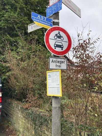 Eine Schildersammlung an einem Pfahl in einem Dorf. Zentral ist das Verbotsschild für Autos und Motorräder angebracht. Darunter befinden sich das Schild für  Anlieger frei, darunter ein Fahrplan für den Rufbus. Weiter oben Hinweisschild für den Anruf