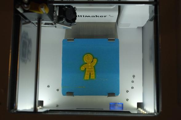 Photograph of a 3D-printed plastic representation of a waving gingerbread man. The figure is on the bed of an Ultimaker 3D printer which is photographed from above. FabLab Zeeland, Middelburg, Netherlands, 6 December 2017. Photo Cindy Kohtala