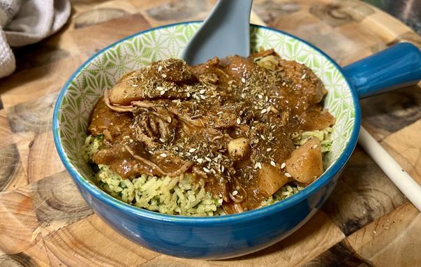 a blue bowl (with a handle) full of food on a wooden butcher block. there is a grey spoon poking out of the food. the food is yellow basmati rice with flecks of spinach, and on top is a rich brown sauce with shredded chicken and chunks of vegetable including potato, topped with a dukkah spice blend.