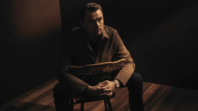 Leonardo DiCaprio assis sur une chaise en bois, les mains croisées, dans une scène à la lumière tamisée et d'atmosphère sombre.
