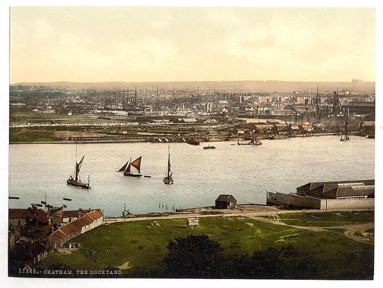 This image is a historical photograph depicting an expansive view of what appears to be the dockyard in Chatham, England. The scene captures several sailing ships with tall masts and distinctive red sails navigating through calm waters that likely indicate a river or harbor setting. In the foreground, there's a well-maintained grassy area leading up to some traditional buildings with pitched roofs, possibly warehouses or administrative structures related to maritime activities.

In contrast to these more modern-looking facilities, in the midground of the image stands an array of larger industrial ships and cranes indicative of active shipbuilding. The background reveals additional infrastructure like docks lined with smaller boats, a network of railway lines likely for transporting goods between various parts of the dockyard or city, and distant buildings that contribute to the urban sprawl.

The overall color palette is dominated by muted earth tones with hints of blue in the water, suggesting an early 20th-century timeframe. The photograph's condition shows some wear indicative of age, such as creases and discoloration typical for prints from this period. These elements collectively paint a picture of bustling industrial activity at Chatham Dockyard during its heyday.