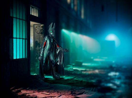 A neoprene clad latex cloaked Jackal appears from a doorway before you, within the decaying remains of an abandoned warehouse. Its dimly lit by cold white, green and blue lighting, giving a very surreal atmosphere to the place. His arm is outstretched as if to get your attention.