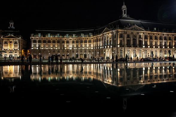Illuminated classical building at night with reflection on water surface, and silhouettes of people in the foreground.