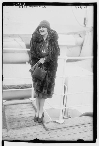The image is a black-and-white photograph of a woman standing on a ship's deck. She is wearing a stylish, dark fur coat with a hood and a fur-lined collar. Underneath the coat, she has a lighter-colored outfit that appears to be a dress with a patterned scarf or sash. She is also wearing dark-colored high-heeled shoes and holding a small, dark purse in her right hand. Her left hand is slightly raised, possibly adjusting her coat or holding something. The woman is smiling and looking to the side, giving a sense of casual elegance. The ship's deck has white railings, and the background shows parts of the ship's structure, including what looks like a lifeboat or part of the ship's equipment. The photograph has a caption at the top that reads "EDNA PURVIANCE," and a number "6481-2" is written on the right side of the image. The overall tone of the image suggests it is from the early 20th century, based on the fashion and photographic style.